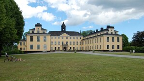 A large, yellow building with a central clock tower, surrounded by a grassy lawn and trees. - Hotell Mörby AB, Patienthotell (Danderyd)