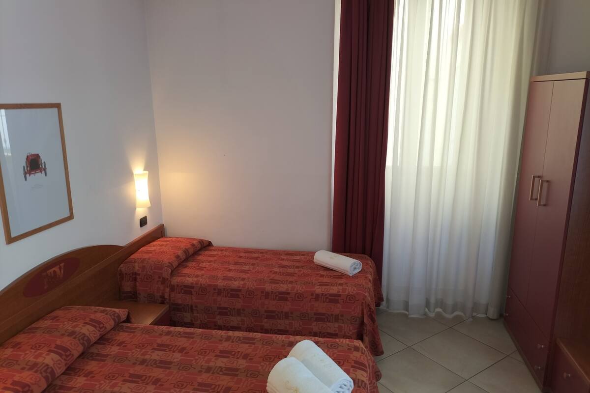 A hotel room with two beds, a wardrobe, a framed picture on the wall, and a window with curtains.