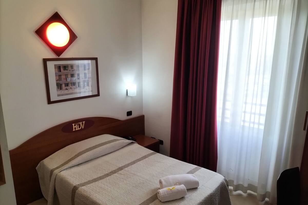 A hotel room with a bed, a headboard with 'HDV' letters, a framed picture on the wall, a nightstand, and a window with curtains.
