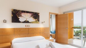 A hotel room with a large bed, a wooden headboard, a painting of a flower, a balcony with a view, and a wooden door. - Bluesea Salina (Santa Eulalia del Rio)