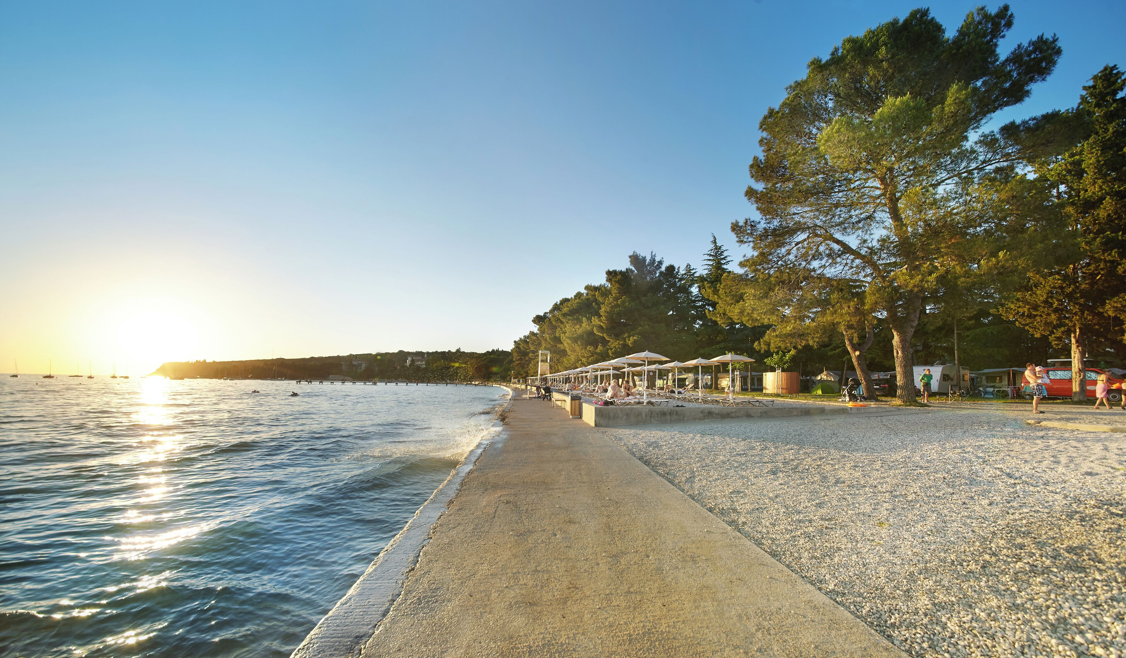 A beach with a paved walkway, umbrellas, and trees.
