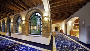 Un pasillo con arcos de piedra, alfombra azul y vista a un espacio exterior con un árbol y un edificio.