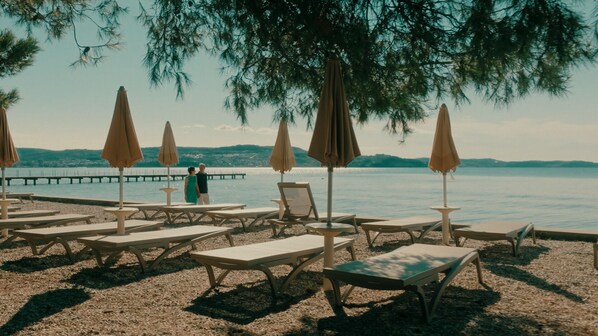 A beachside lounge area with multiple sun loungers and umbrellas, a pier in the background, and a couple standing by the water.