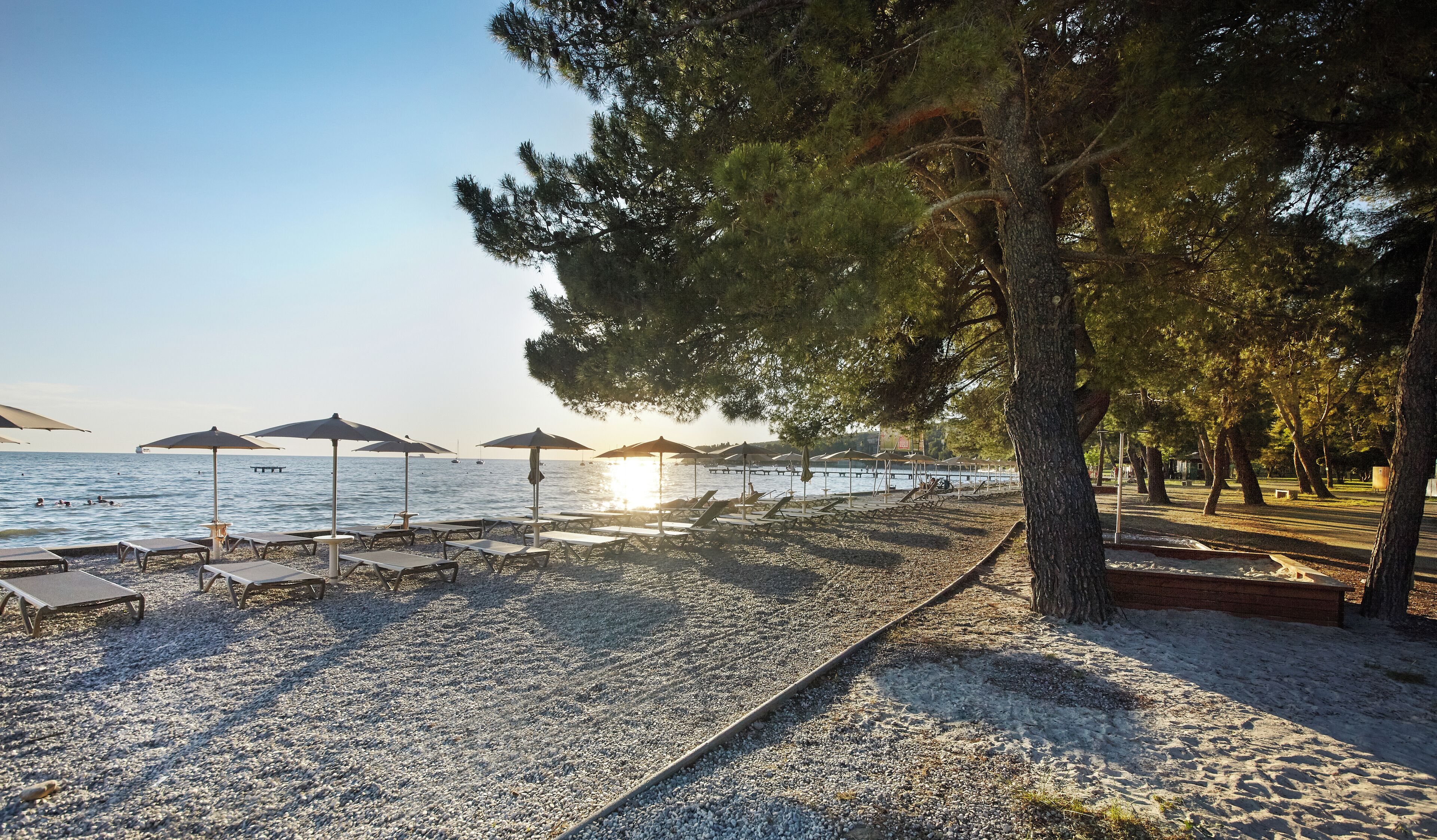 Ein Kiesstrand mit Liegestühlen und Sonnenschirmen, ein klarer Himmel und ein ruhiges Meer.