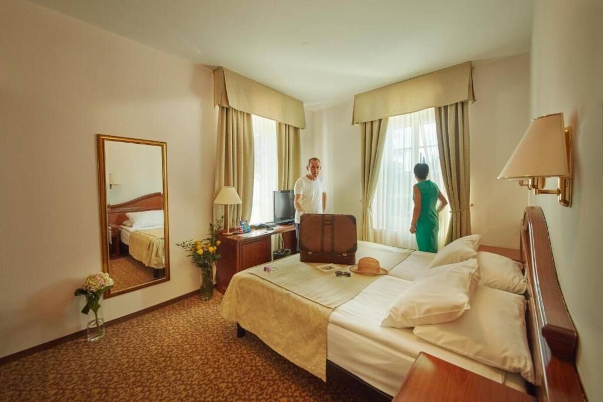 A hotel room with a bed, a desk with a lamp, a mirror, and a window with curtains.