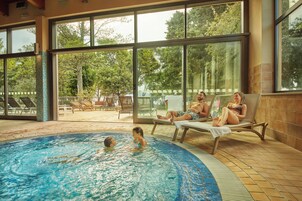 Una piscina interior con paredes de vidrio transparente, sillones de descanso y vistas a árboles.