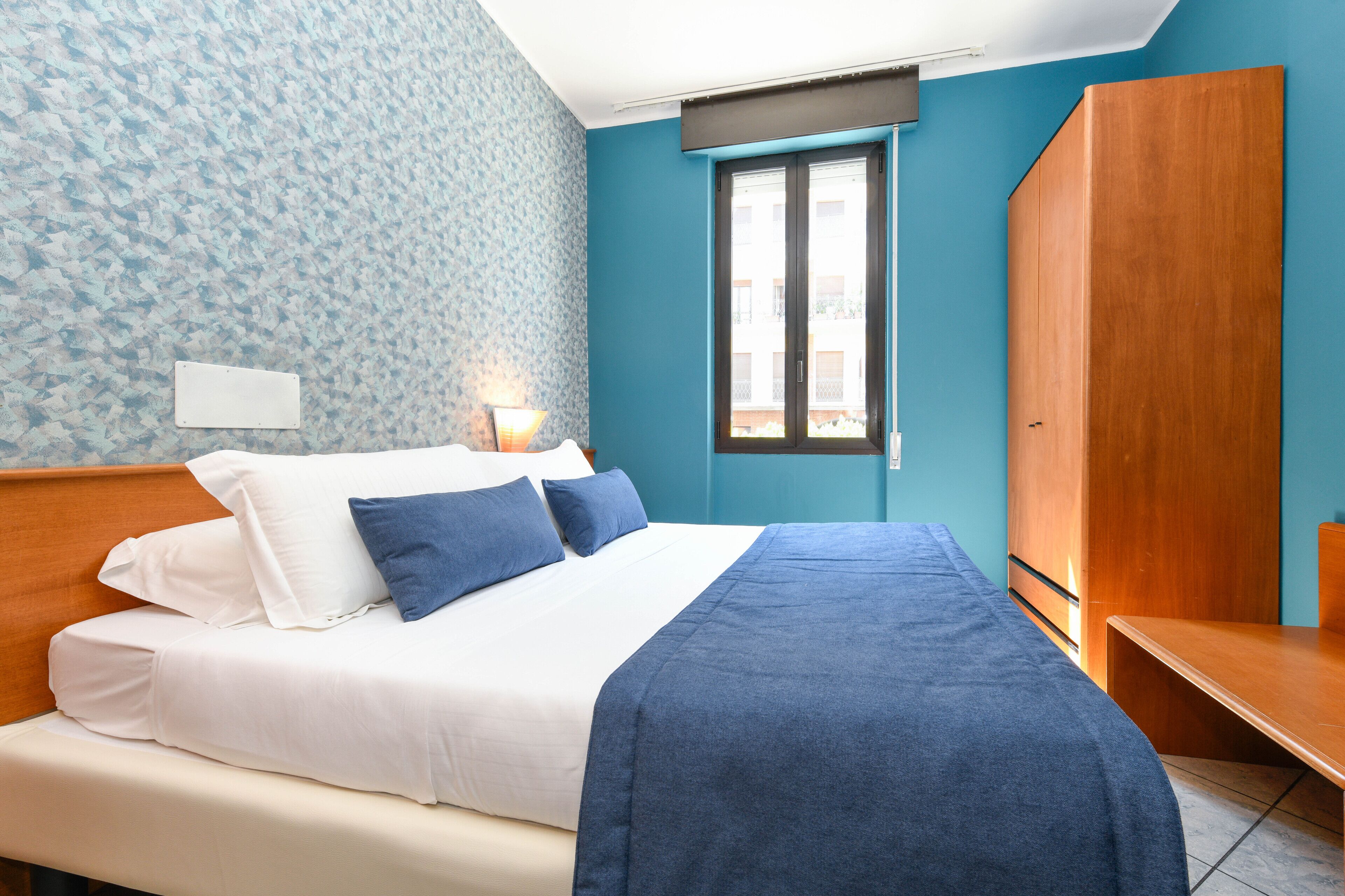 Ein Schlafzimmer mit einem Bett, einem Holzschrank, einem Fenster und einer Wand mit ornamentalem Wandpapier.