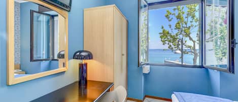Una habitación con escritorio de madera, una silla, una cama, un armario, televisor montado en la pared y ventana con vista al mar.