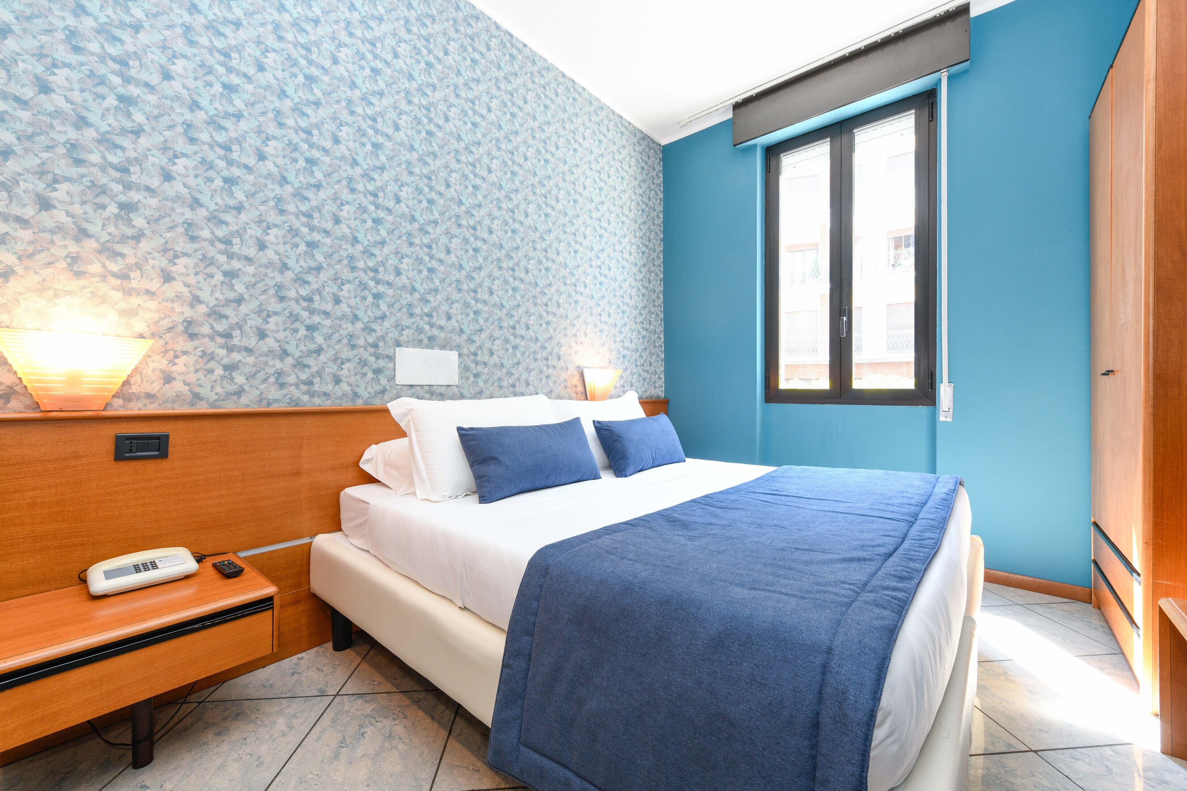 Una habitación de hotel con cama, mesita de noche, lámpara y ventana con persianas.