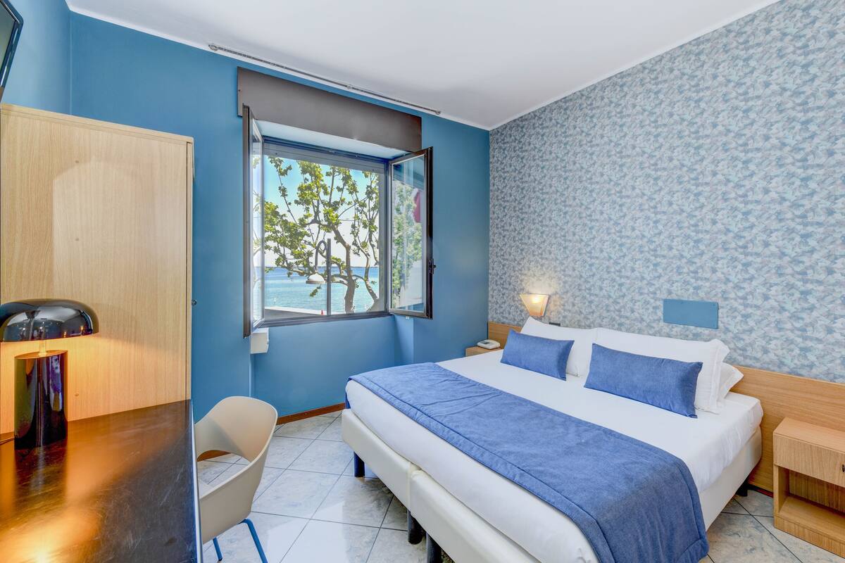 A bedroom with a bed, a desk, a chair, a window with a view of trees and the sea, and a patterned wall.