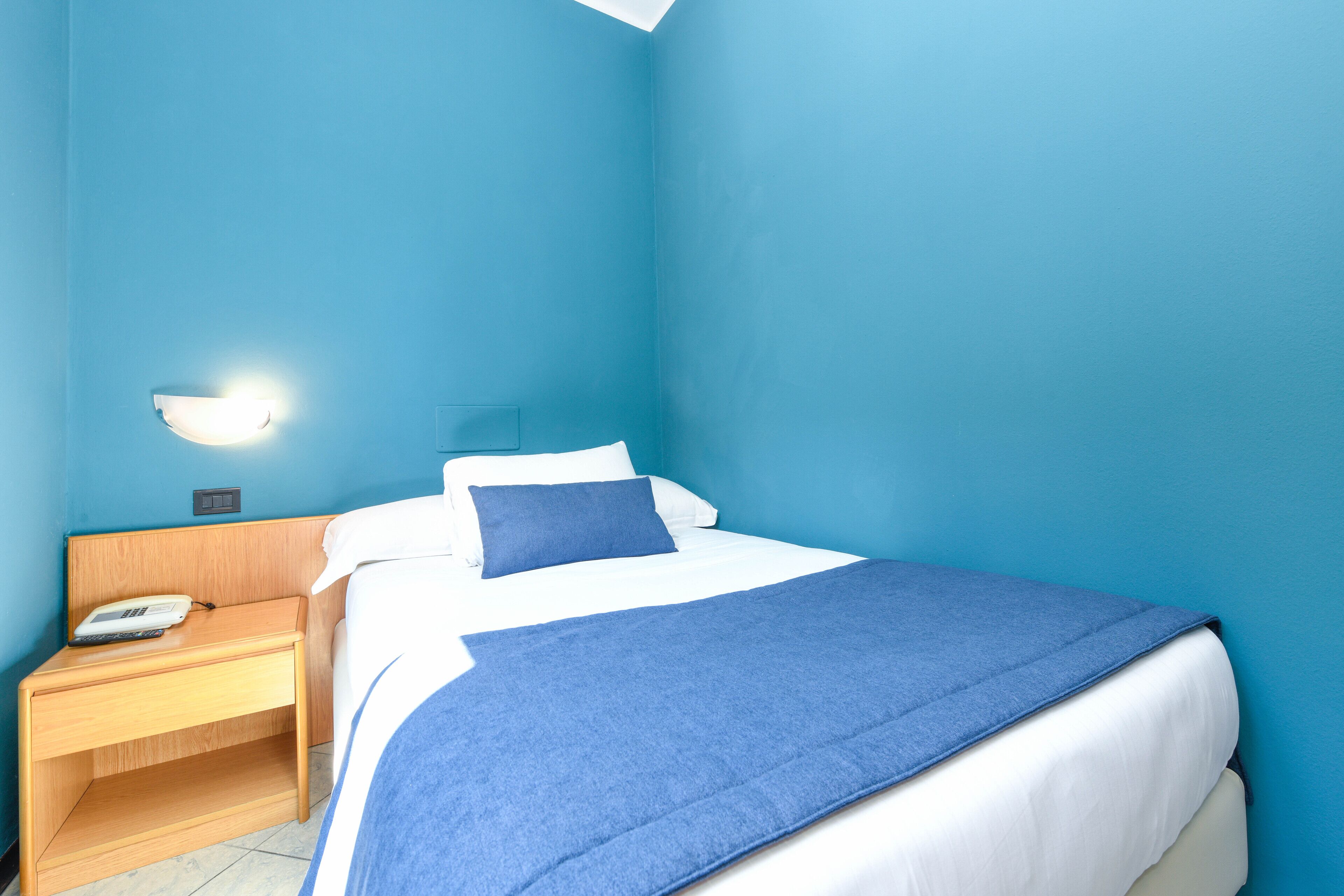 A room with a bed, a nightstand, a wall-mounted light, and a power outlet.
