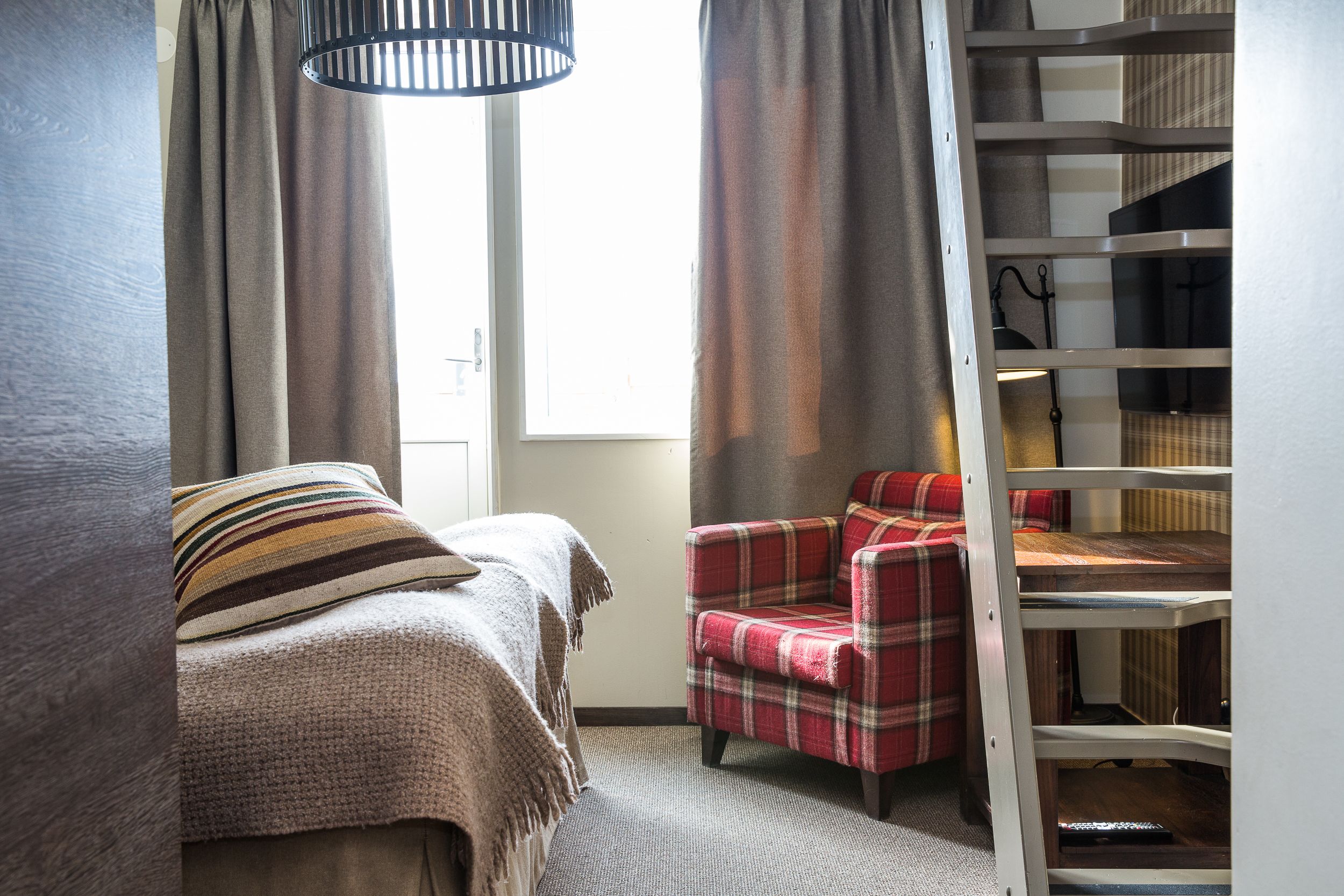 Une chambre à coucher avec un lit, un fauteuil à motifs écossais, une étagère en bois et une fenêtre avec des rideaux.