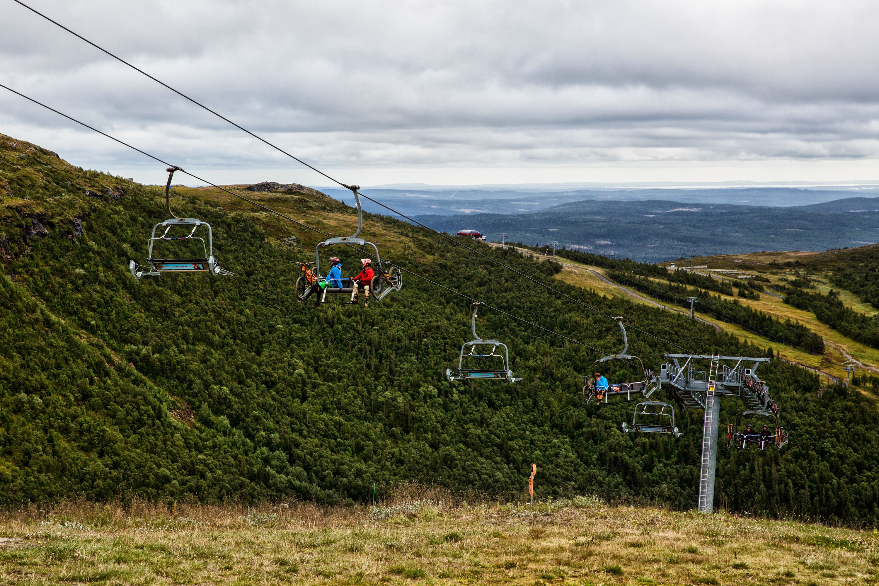 A ski lift with passengers ascending a green, forested mountain slope.