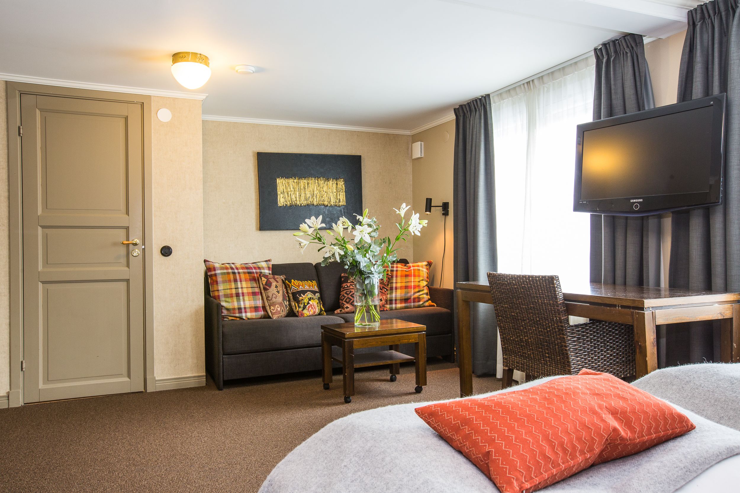 A hotel room with a sofa, a small table with a vase, a television, a desk, and a chair.