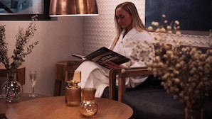 A woman reading a magazine in a cozy, well-lit room with a large window, a round wooden table, and a vase of flowers.