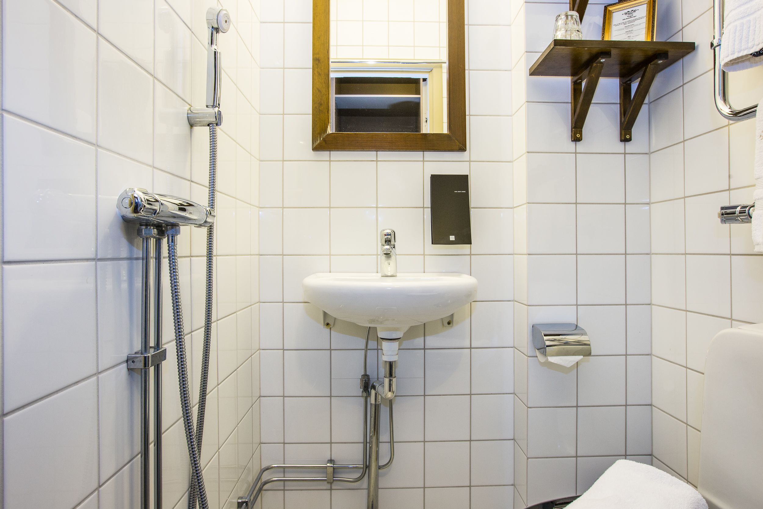 A bathroom with white tiled walls, a white sink, a handheld showerhead, a towel rack, and a small shelf with a framed certificate.