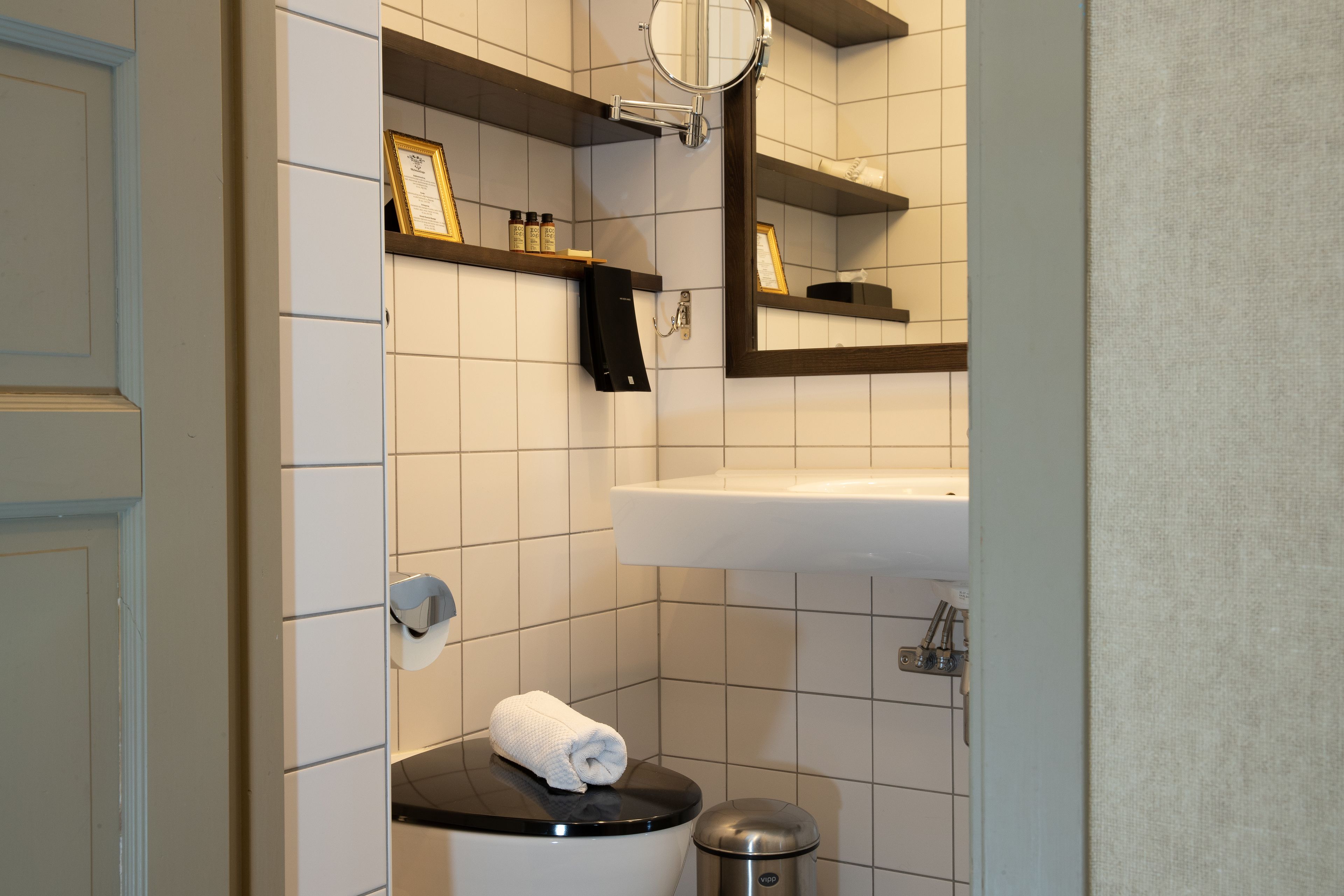 Uma casa de banho moderna com sanita, lavatório e espelho. Existem prateleiras com produtos de higiene pessoal e uma toalha na sanita.