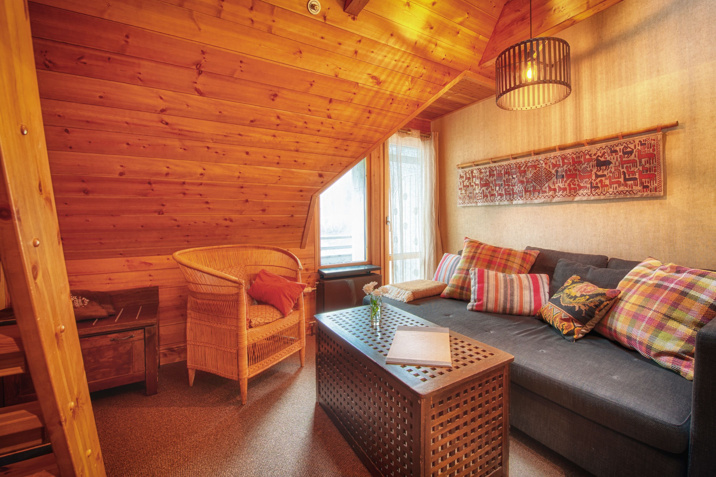 Una habitación acogedora con techo de madera, un sofá con cojines coloridos, una silla de mimbre y una mesita con un libro y flores.