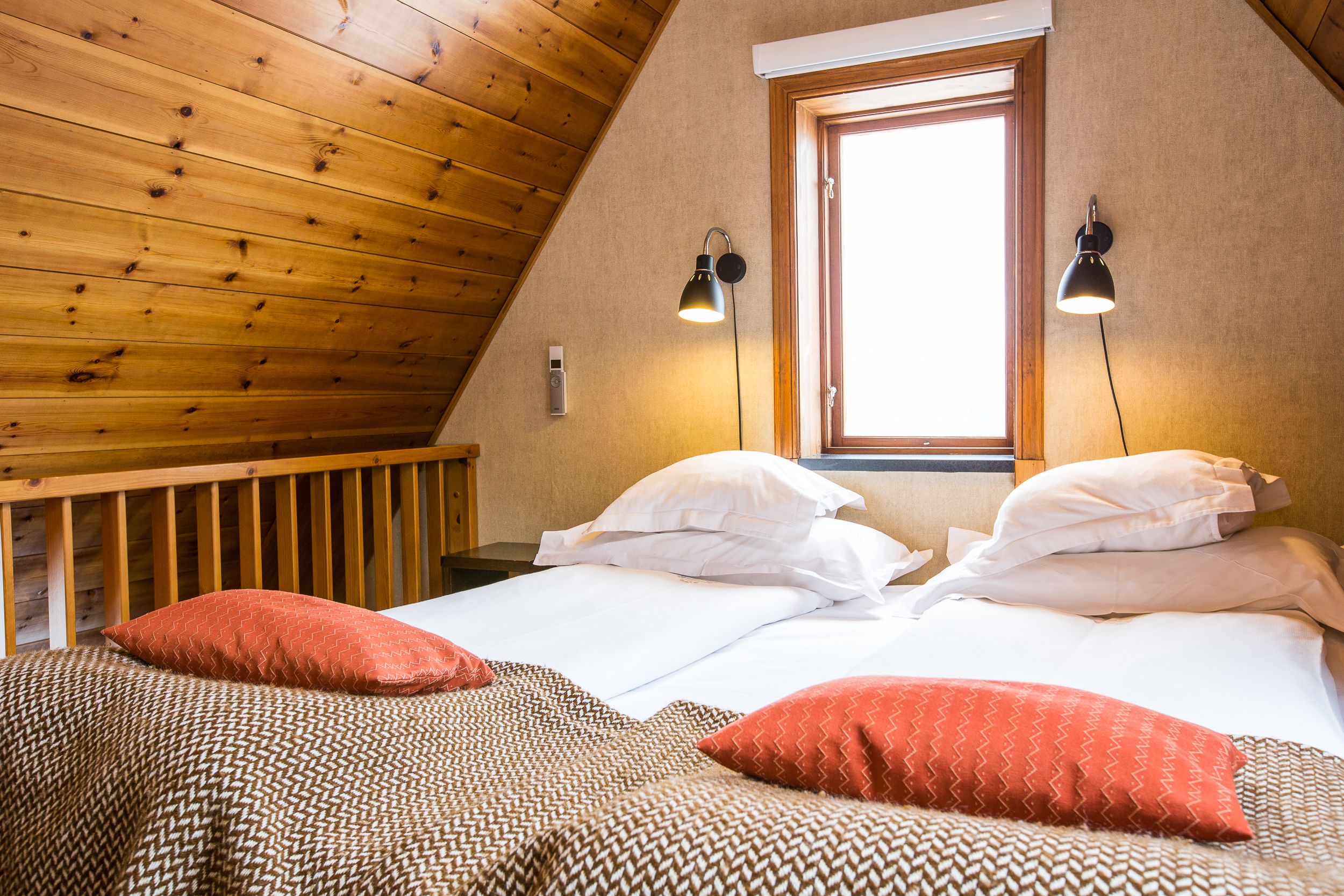 A cozy bedroom with a sloped ceiling, wooden walls, two bedside lamps, a window, and a bed with patterned bedding and pillows.