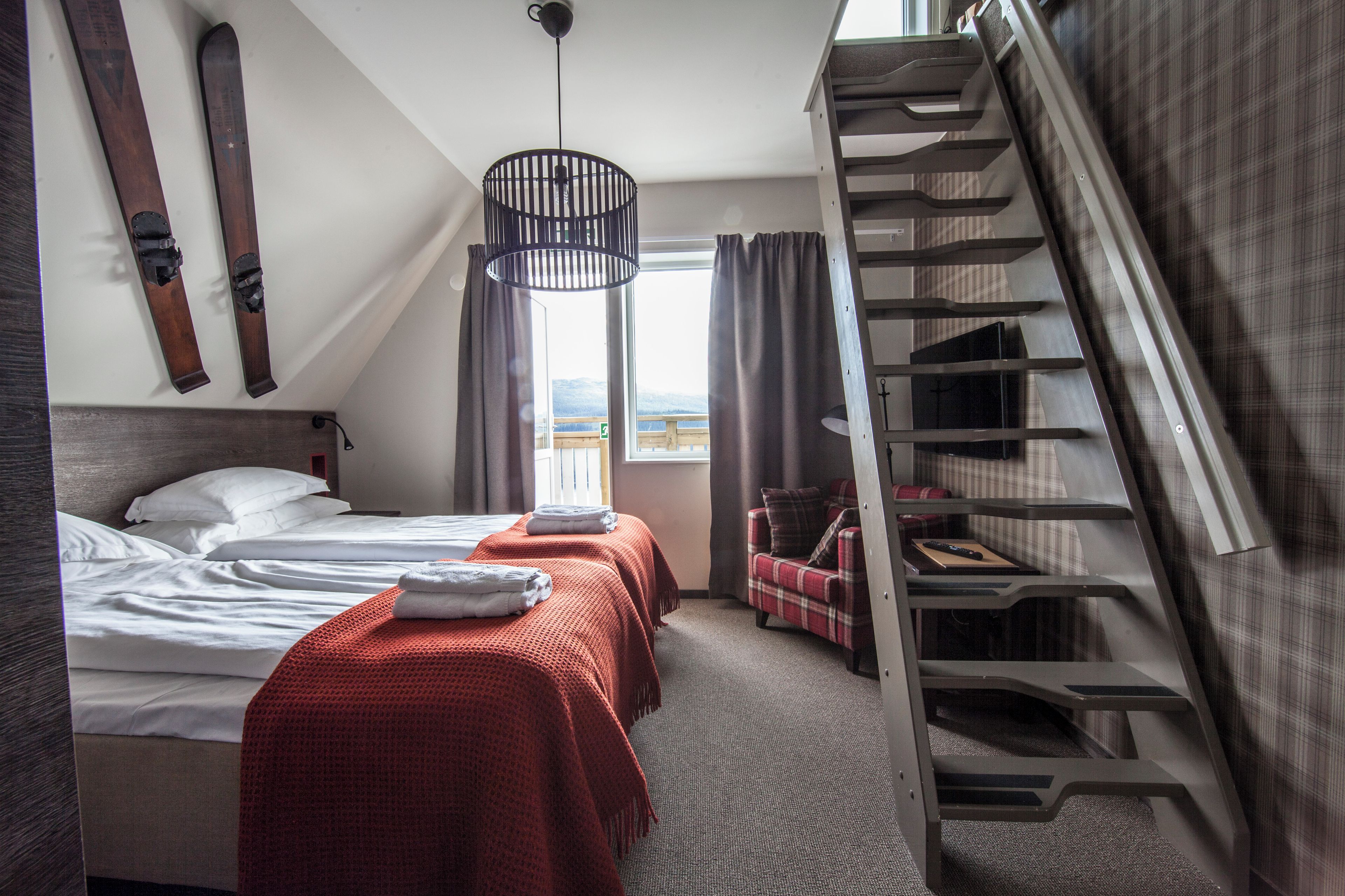 A hotel room with two beds, a red blanket, a wooden headboard, a window with curtains, a view of the mountains, and a staircase.