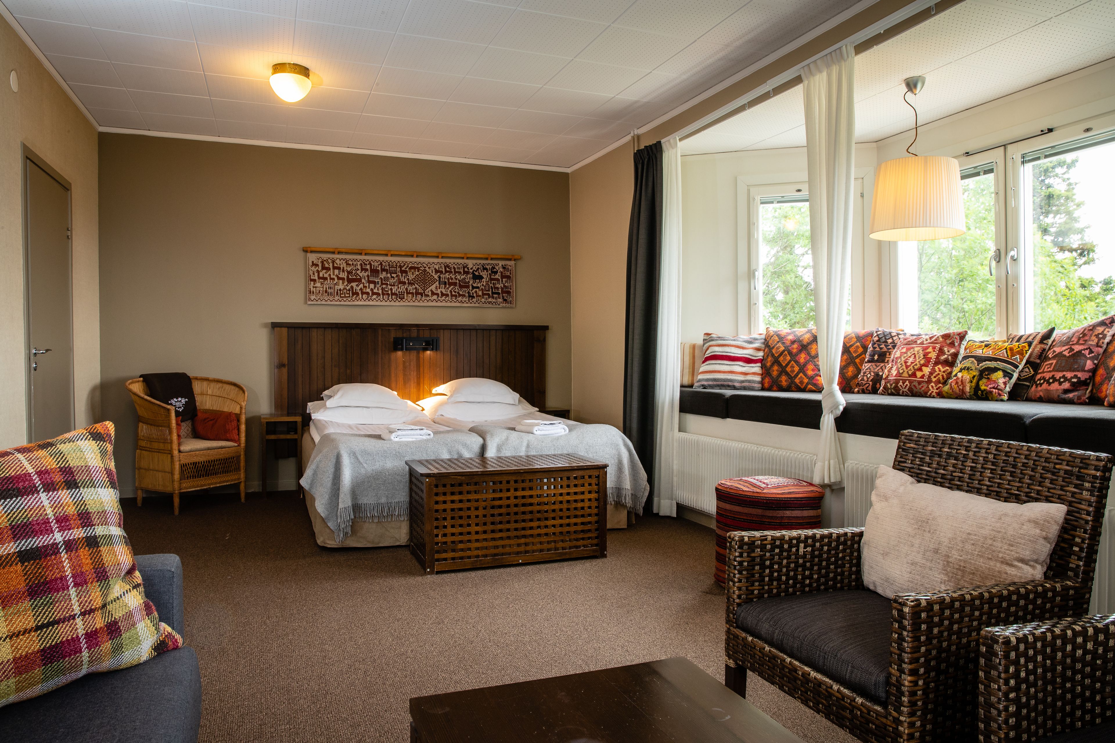 A cozy hotel room with a bed, two wicker chairs, a small table, and a window with a view of greenery.