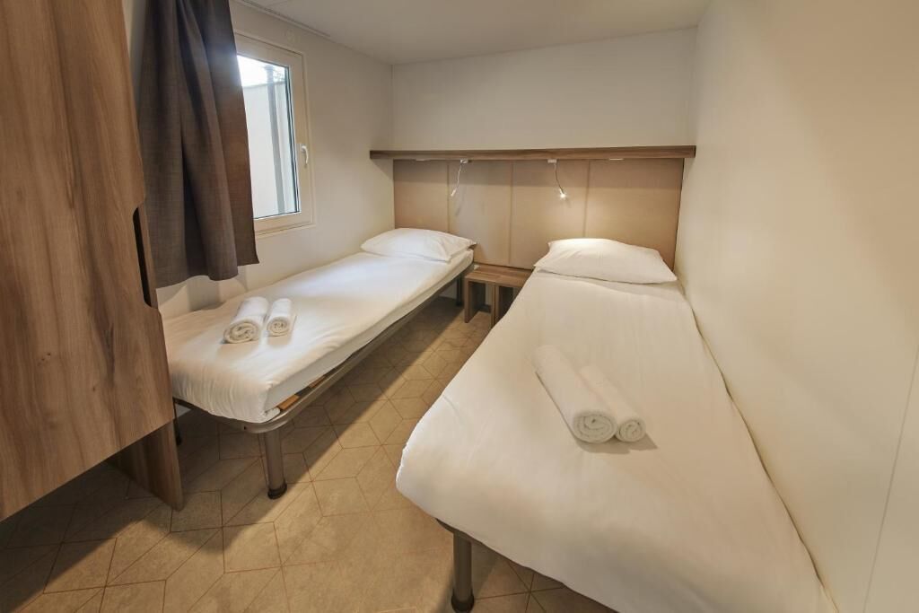 A small, clean room with two single beds, a wooden headboard, a window, and a curtain.
