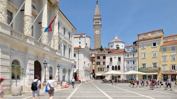 Una amplia plaza con una destacada torre del reloj al fondo, rodeada de edificios históricos con fachadas ornamentales y un cielo azul despejado.