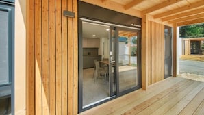 Een houten gevel met een glazen voordeur, een overdekt terras en een zichtbare keukenruimte binnen.