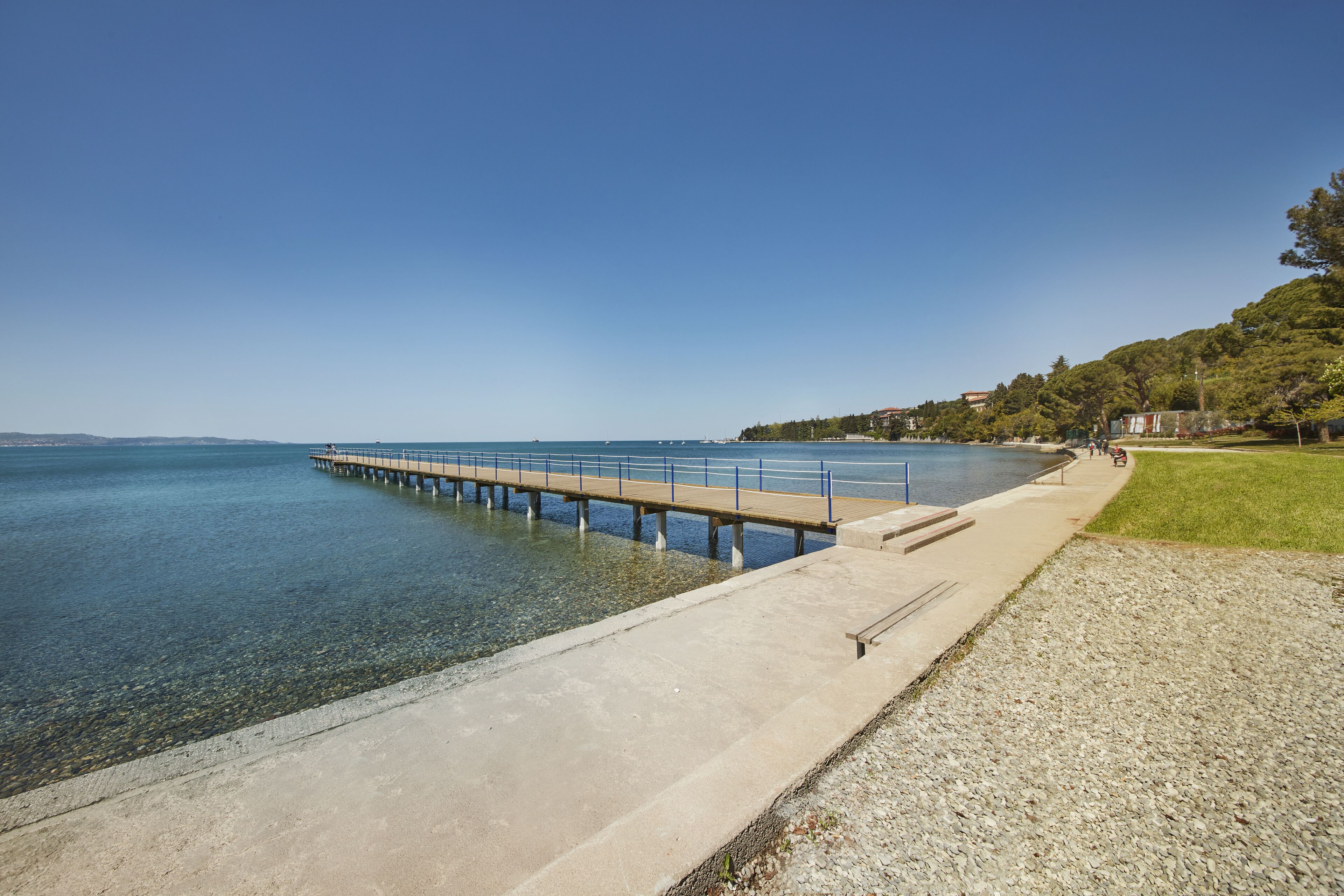 A pier extending into calm waters, a clear sky, and a grassy area with trees.