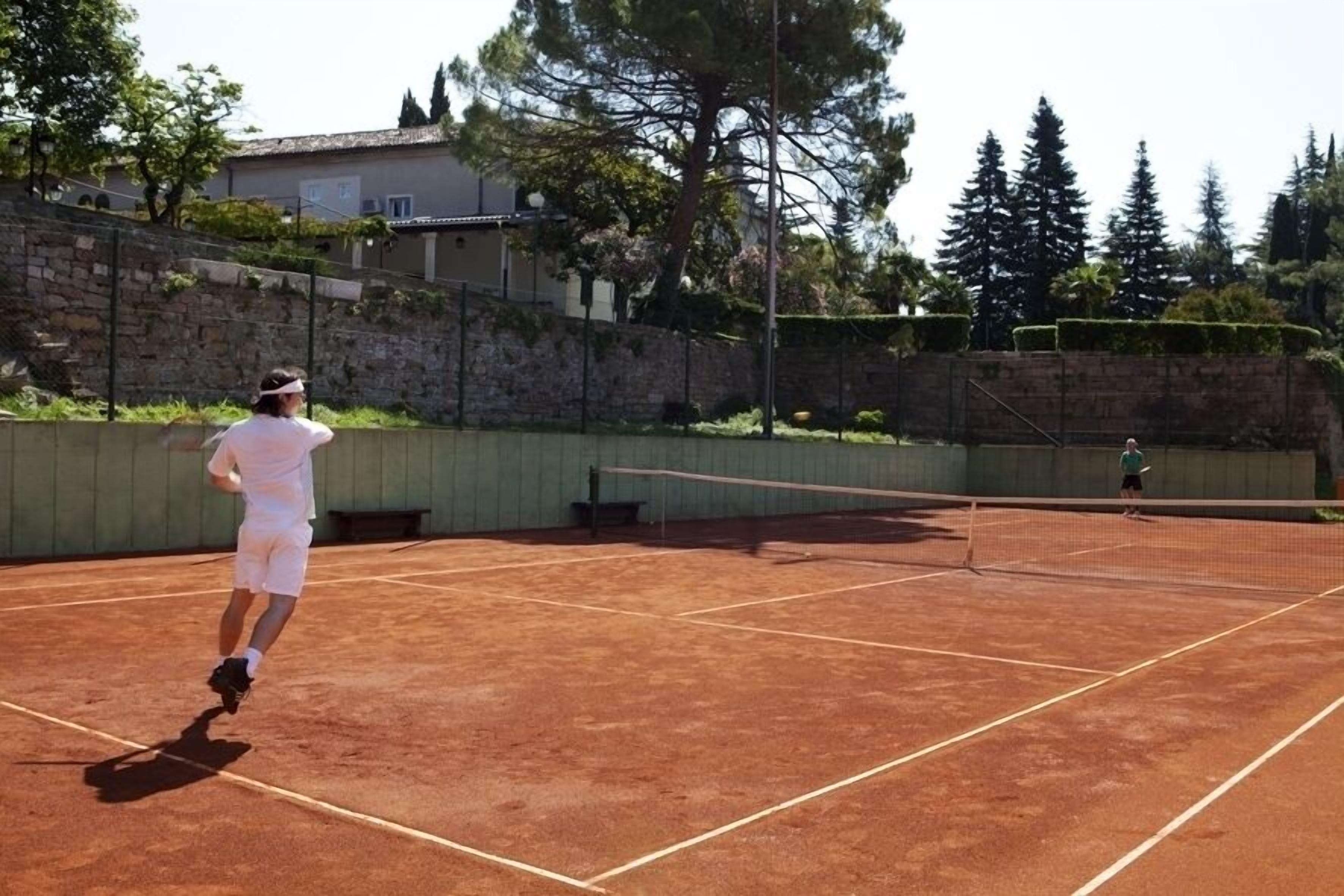 A tennis court with a net, surrounded by a stone wall and trees.