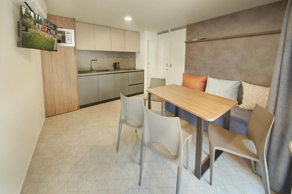 Un espace de vie compact comprenant un coin cuisine, un espace repas avec une table et des chaises en bois, et un lit avec des oreillers.