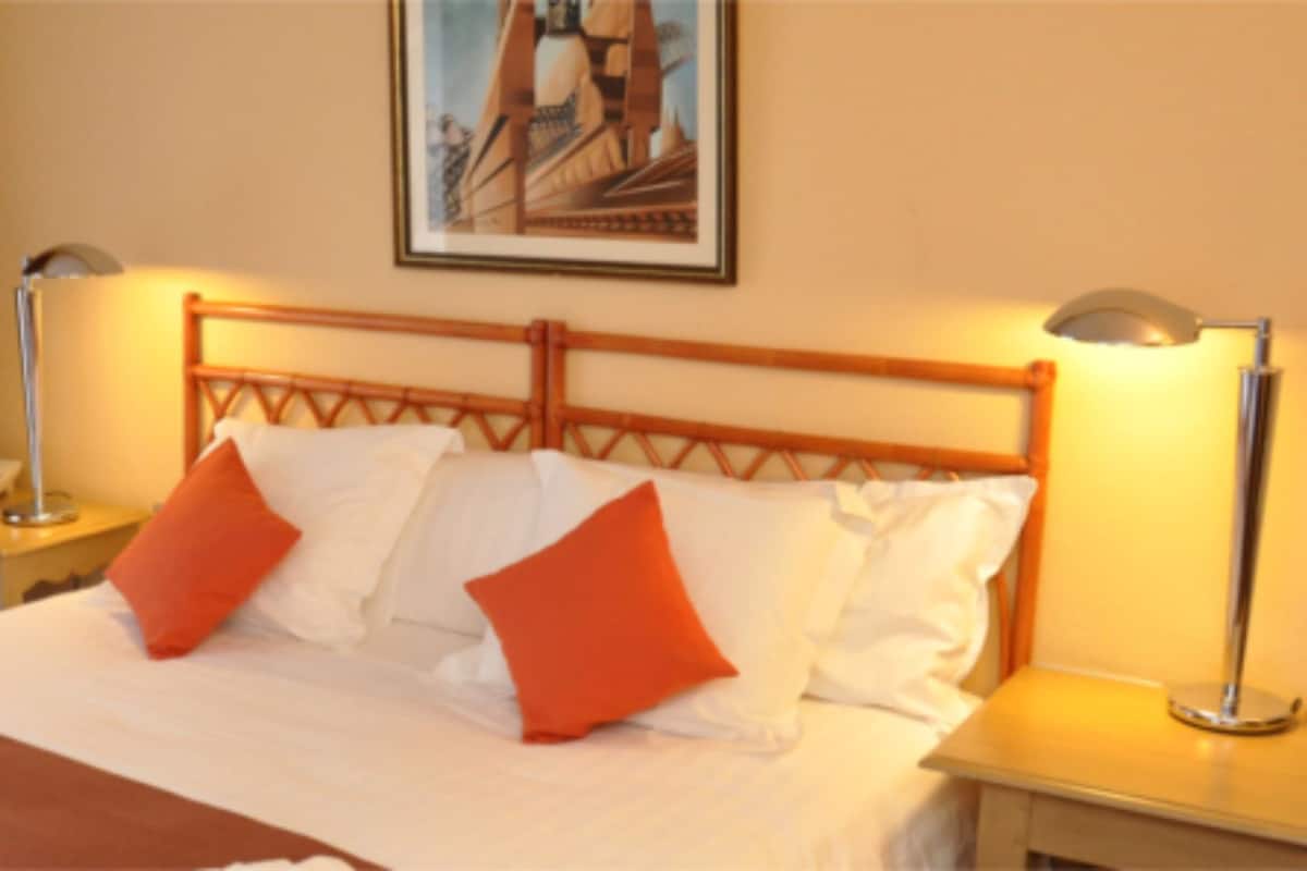 Quarto de hotel com uma cama, duas mesas de cabeceira, uma luminária, um quadro na parede e uma janela com cortinas.