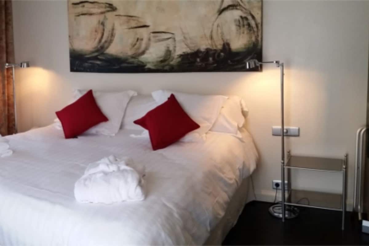 Cama bem arrumada com almofadas vermelhas, um cobertor branco e uma toalha ao lado. Há um quadro na parede acima da cama e um abajur ao lado.