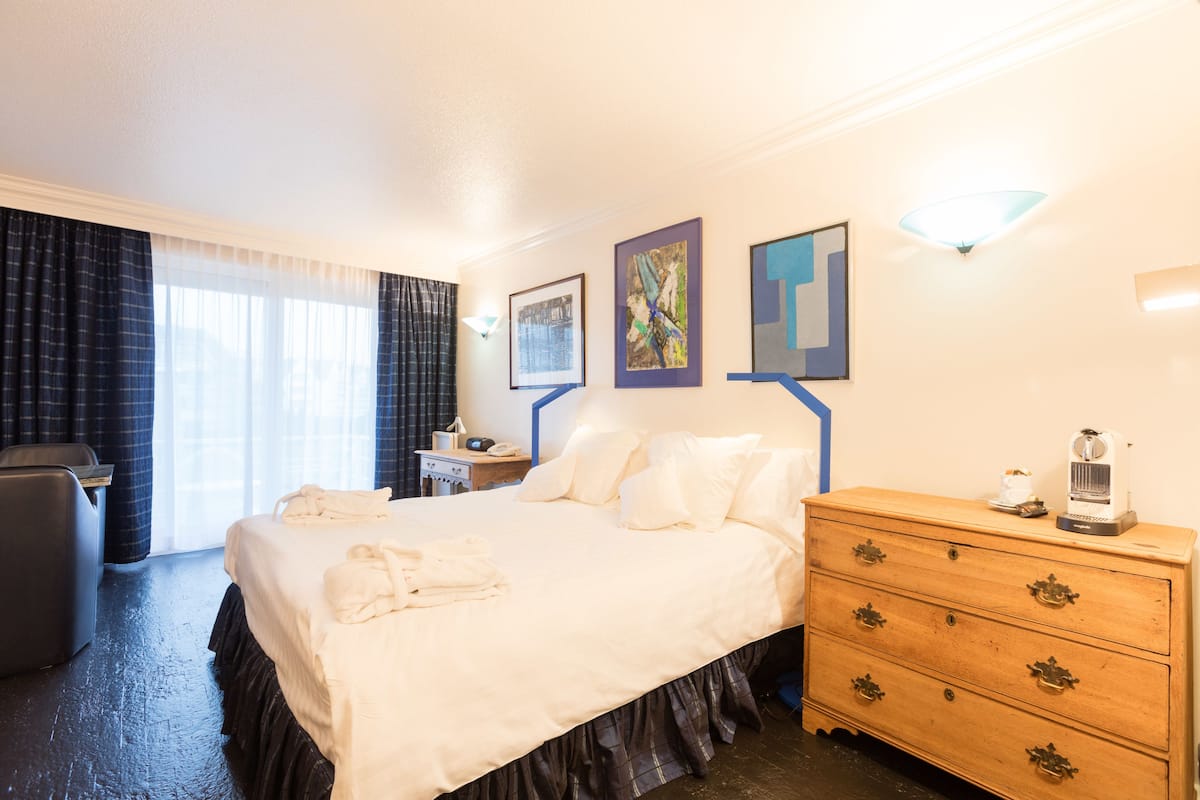 Quarto de hotel com uma cama grande, penteadeira de madeira, escrivaninha com luminária e obras de arte nas paredes.