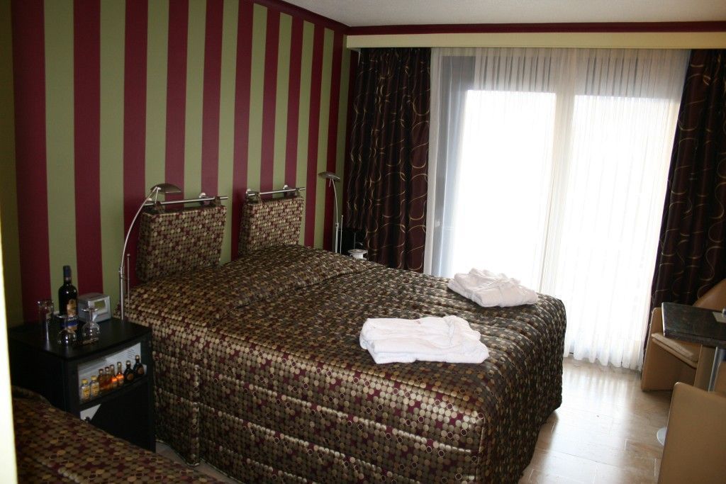 Ein Hotelzimmer mit einem großen Bett, einem Nachttisch mit Lampe, einem kleinen Tisch mit Flaschen und einem Fenster mit Vorhängen.