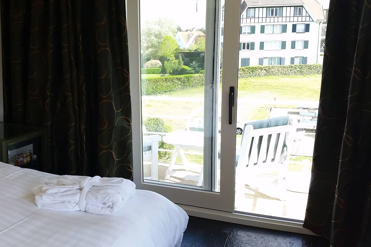 Quarto de hotel com uma cama, dois travesseiros verdes, um abajur, um telefone e uma janela com vista para um edifício e vegetação.
