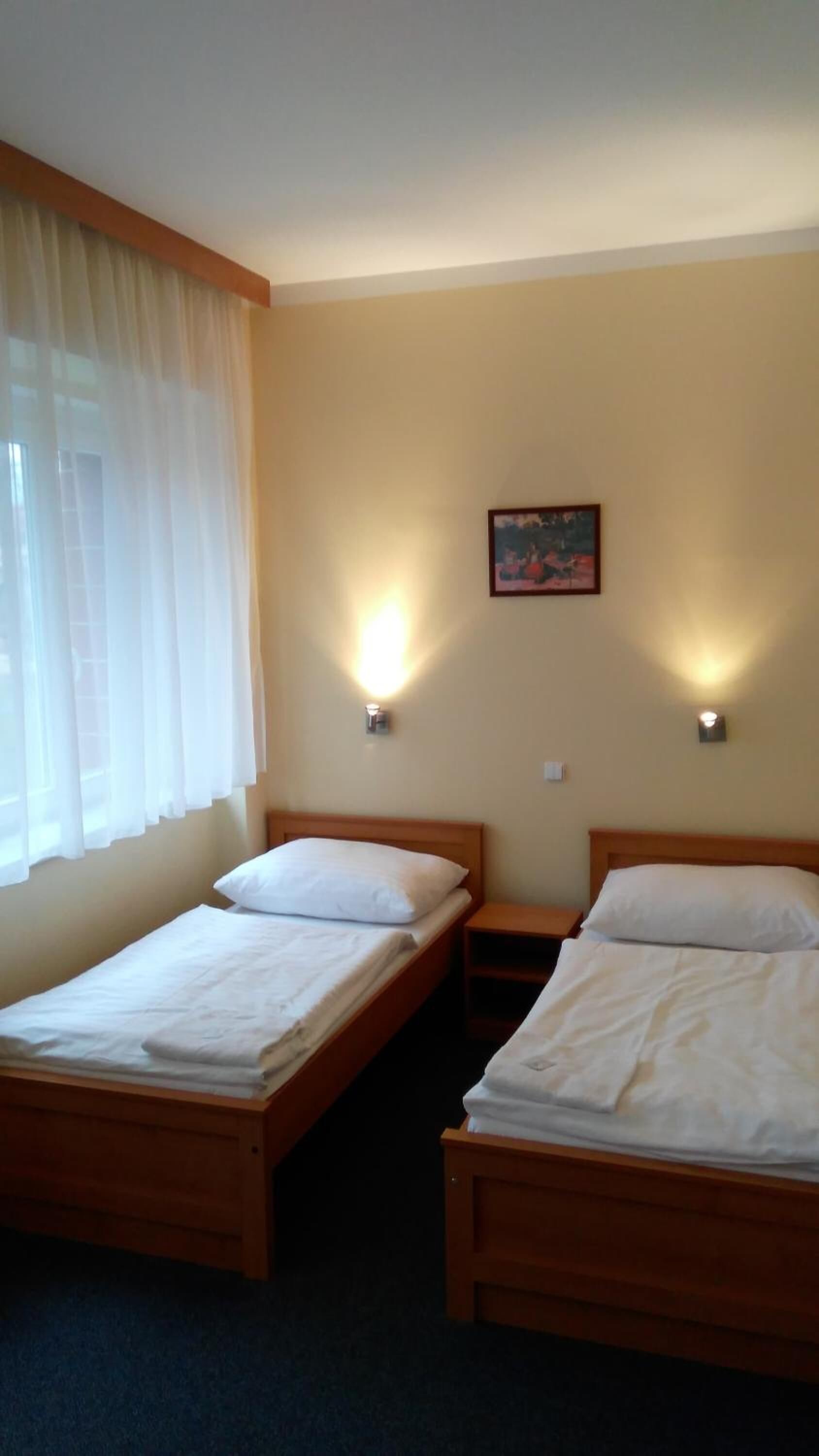 Een hotelkamer met twee enkele bedden, een raam met dunne gordijnen en twee wandlampen.