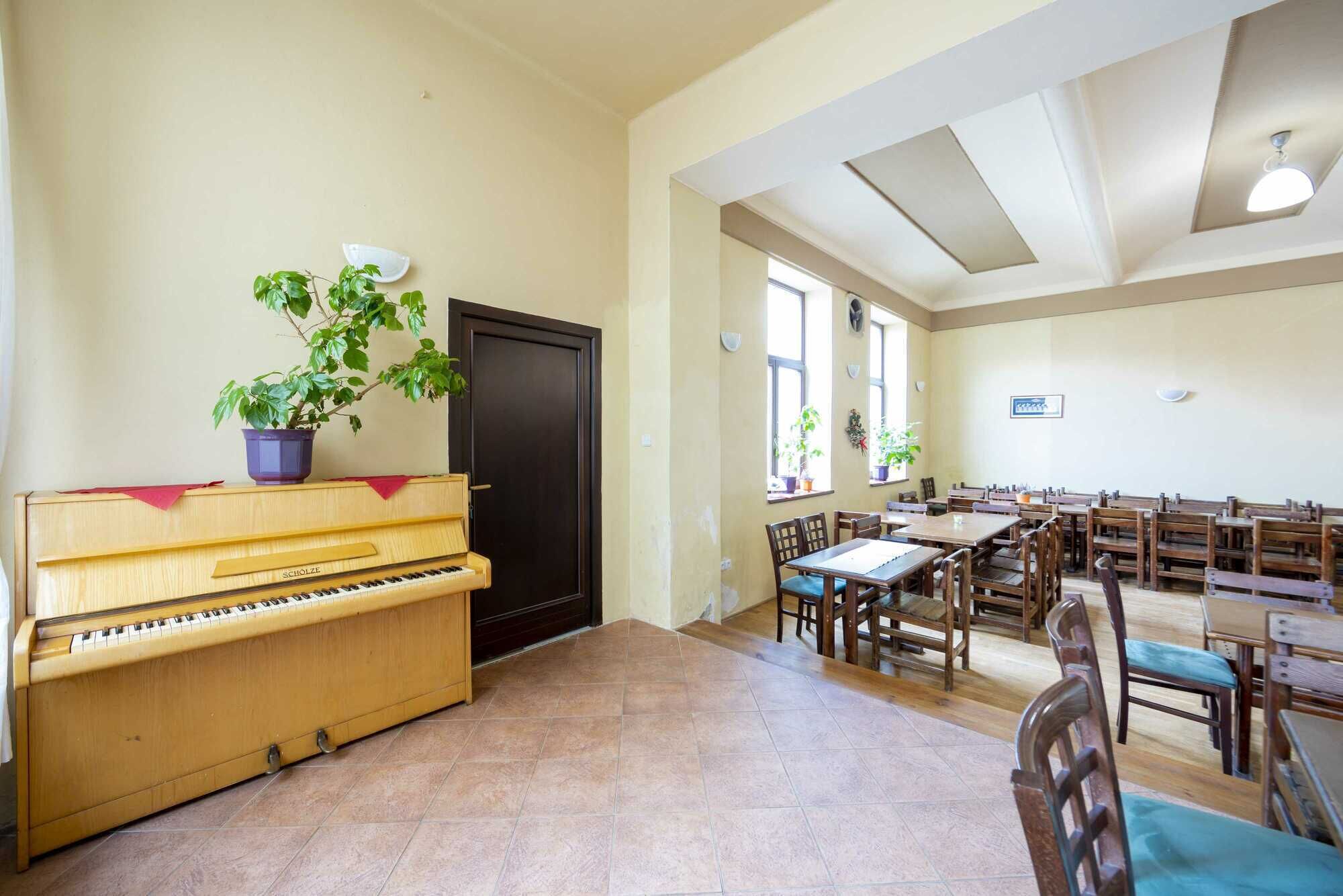 A spacious room with a piano, multiple tables, and chairs, and large windows allowing natural light.