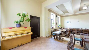 A spacious room with a piano, multiple tables, and chairs, and large windows allowing natural light.