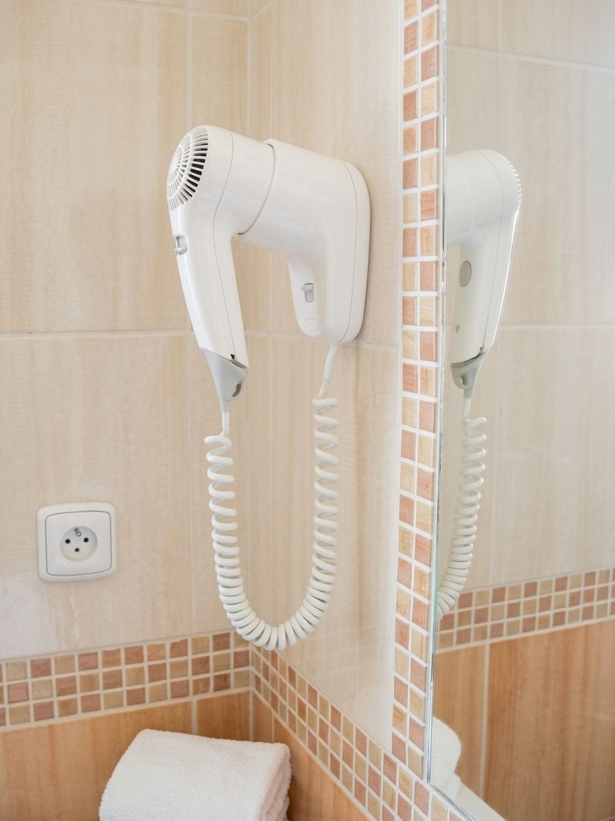A bathroom with two hair dryers mounted on the wall, a towel stacked on the floor, and a tiled shower area.