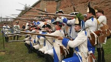 Un grupo de personas vestidas con trajes de época, con largas lanzas de madera, arrodilladas apuntando hacia adelante.