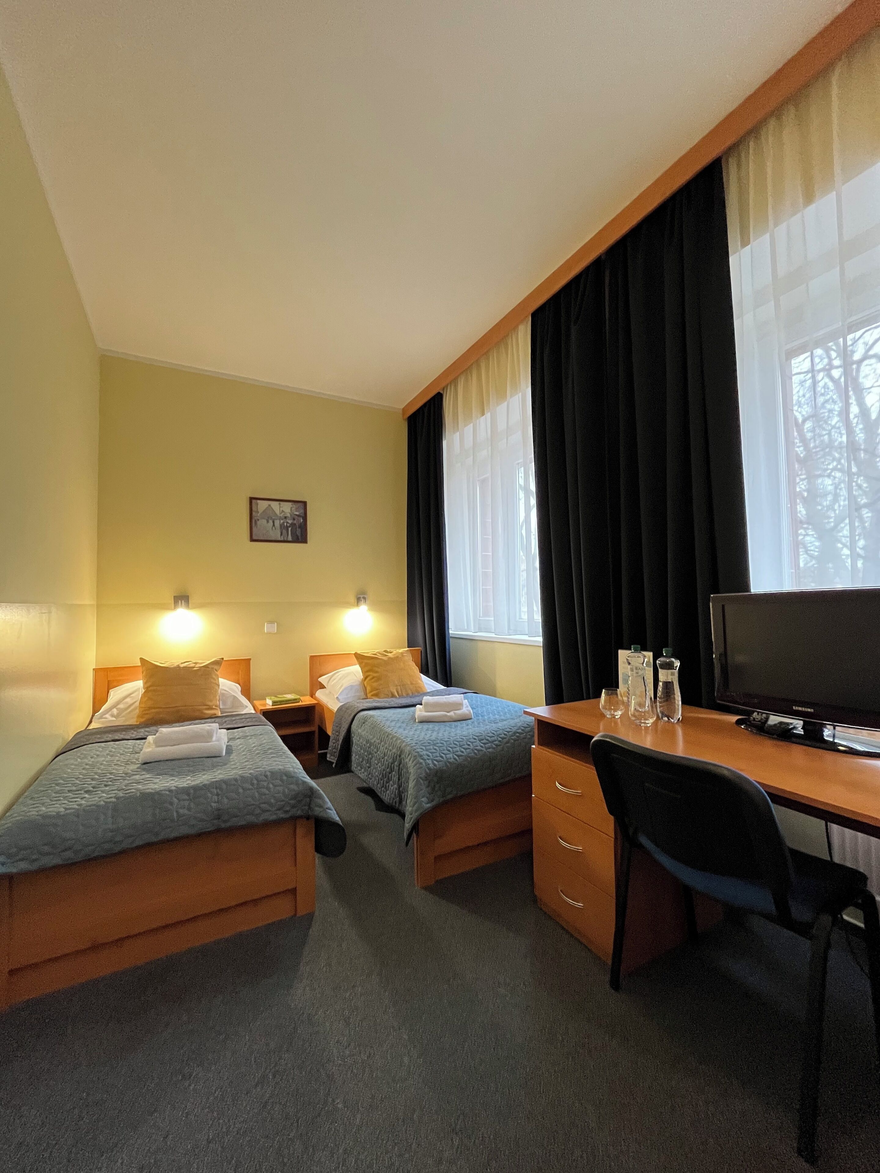 A hotel room with two single beds, a desk with a computer, and a window with curtains.