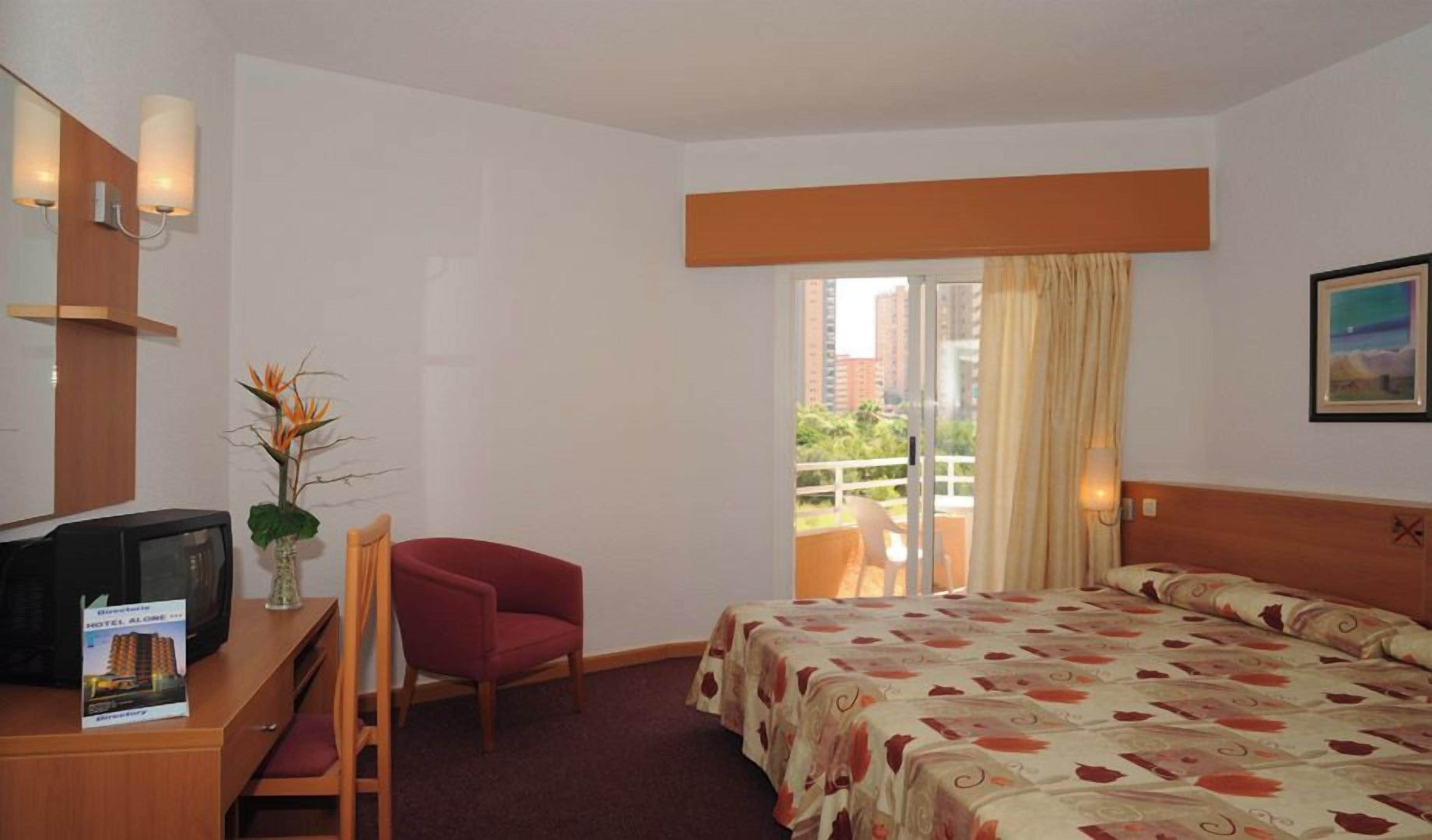 A hotel room with a bed, a desk with a television, a chair, a plant, and a window with a view of buildings.