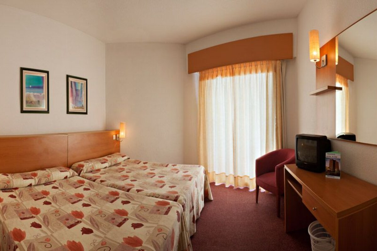 A hotel room with two beds, a desk with a television, a chair, a waste bin, and two framed pictures on the wall.