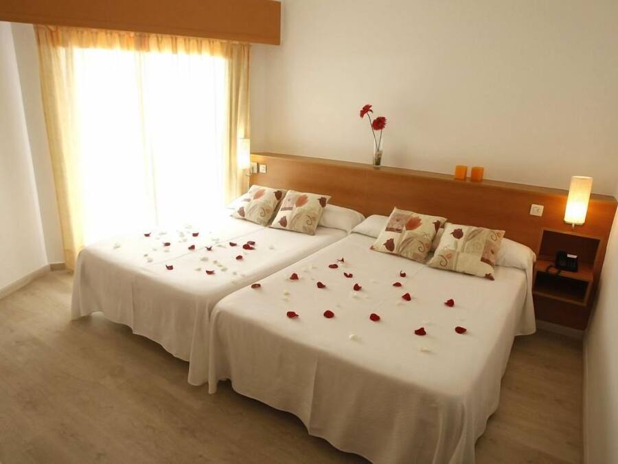 A hotel room with two beds, a nightstand, a lamp, and a window with curtains.