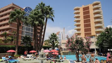 A hotel pool area with multiple levels of buildings, palm trees, and a fountain.