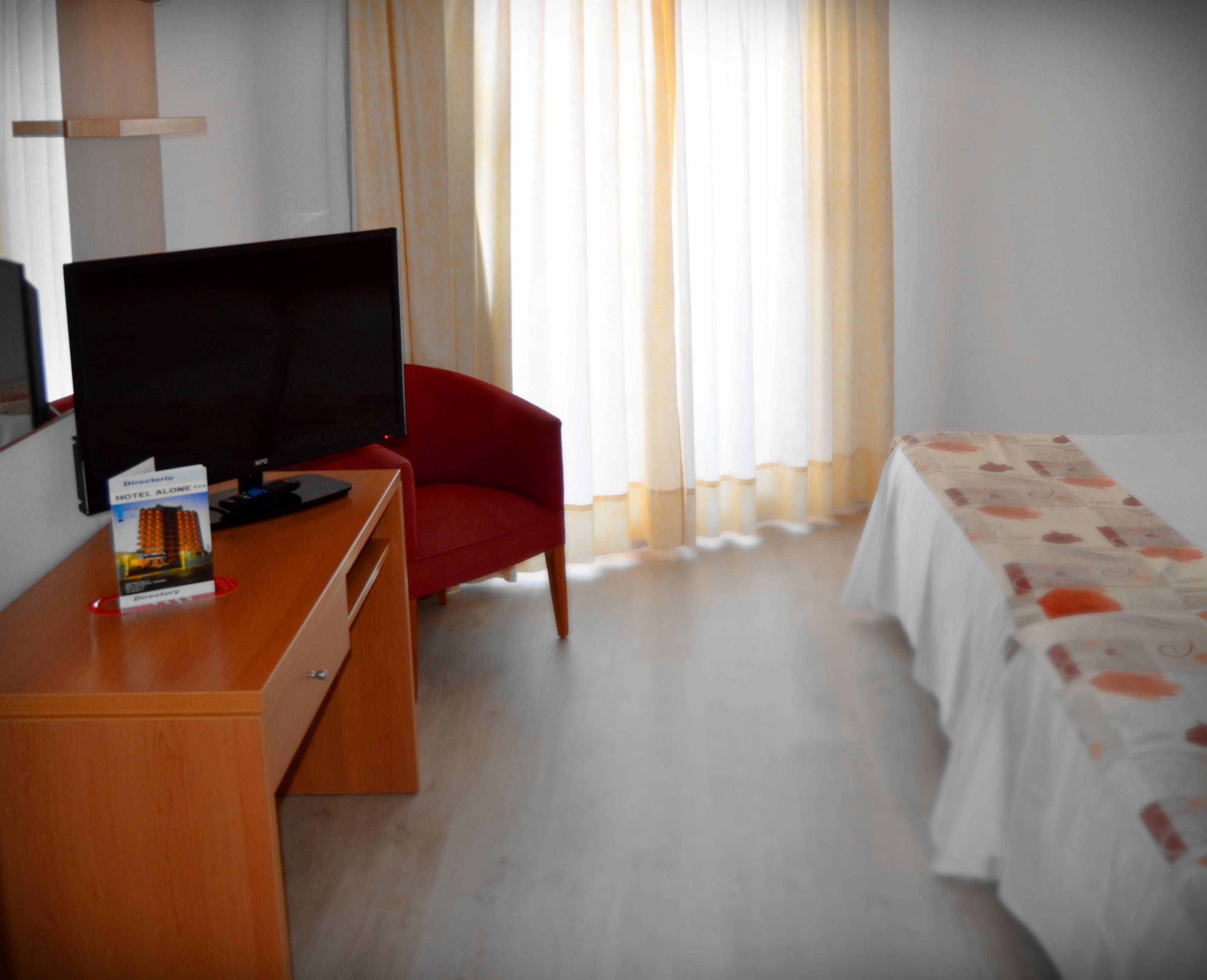A hotel room with a TV on a wooden desk, a red chair, a bed with a white and orange patterned bedspread, and a window with sheer curtains.