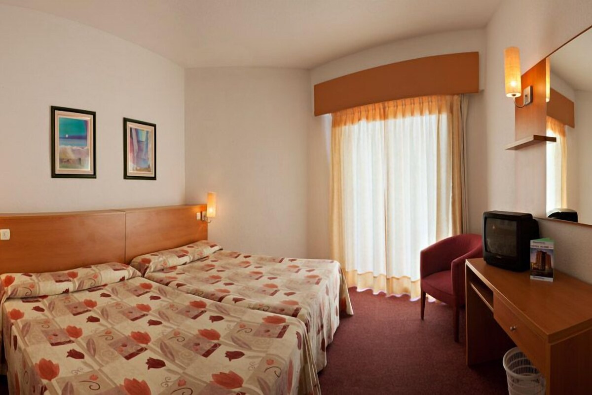 Habitación de hotel con dos camas, un escritorio con televisor, una silla, un cesto de basura y dos cuadros enmarcados en la pared.