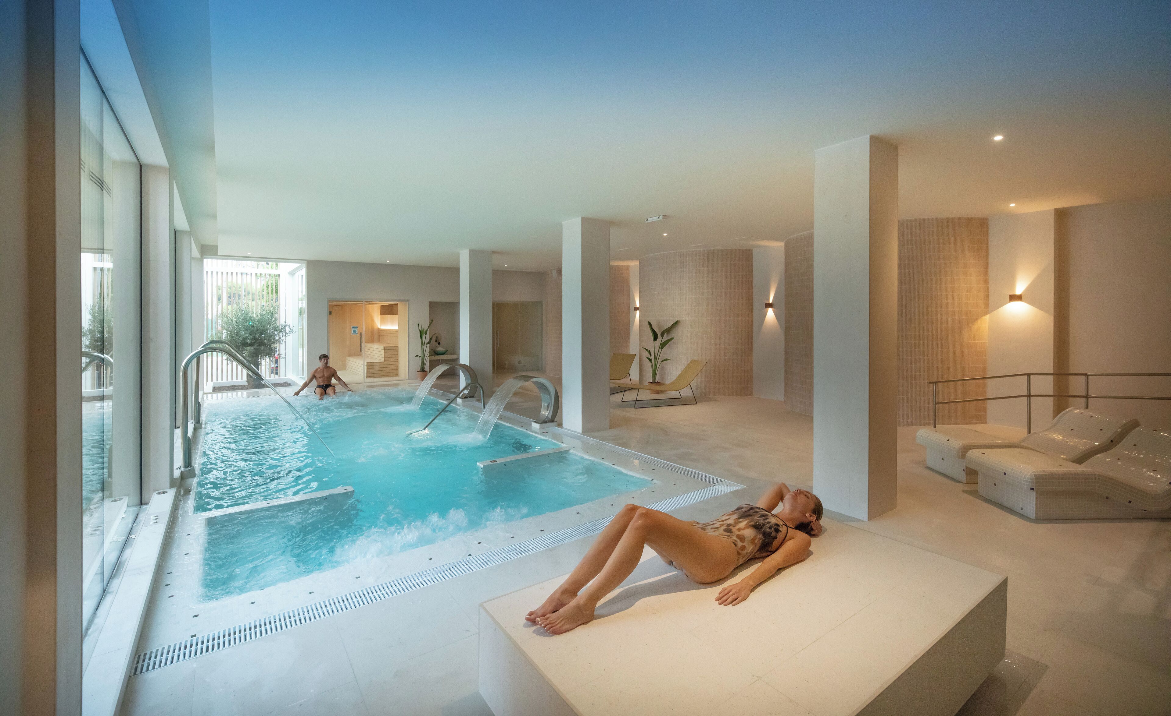 A modern indoor pool area with a person relaxing on a sun lounger, a separate pool with a water slide, and a spa area with chairs and a wall-mounted light.