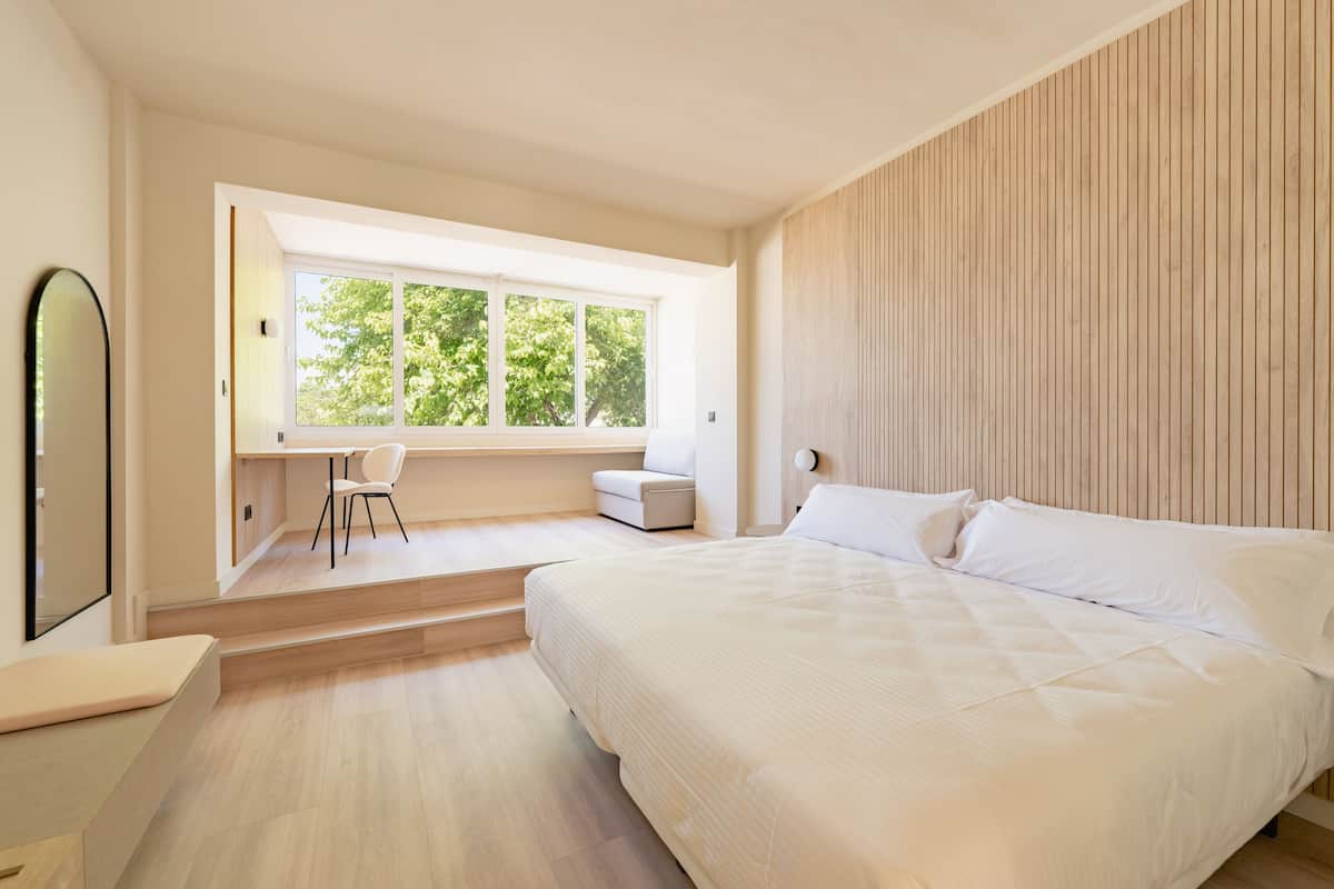 A modern bedroom with a large bed, a desk, and a chair, a window with a view of trees, and a wooden wall.
